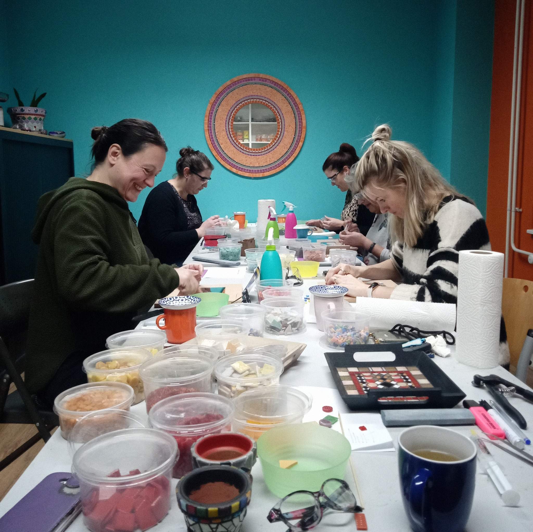 Vier enthousiaste cursisten doen mee met een mozaiekworkshop. De cursisten zitten aan een lange tafel gevuld met mozaiekmateriaal en voorbeelden. De workshopruimte is kleurrijk ingericht met een turquoise muur en een vrolijke mozaiekspiegel.