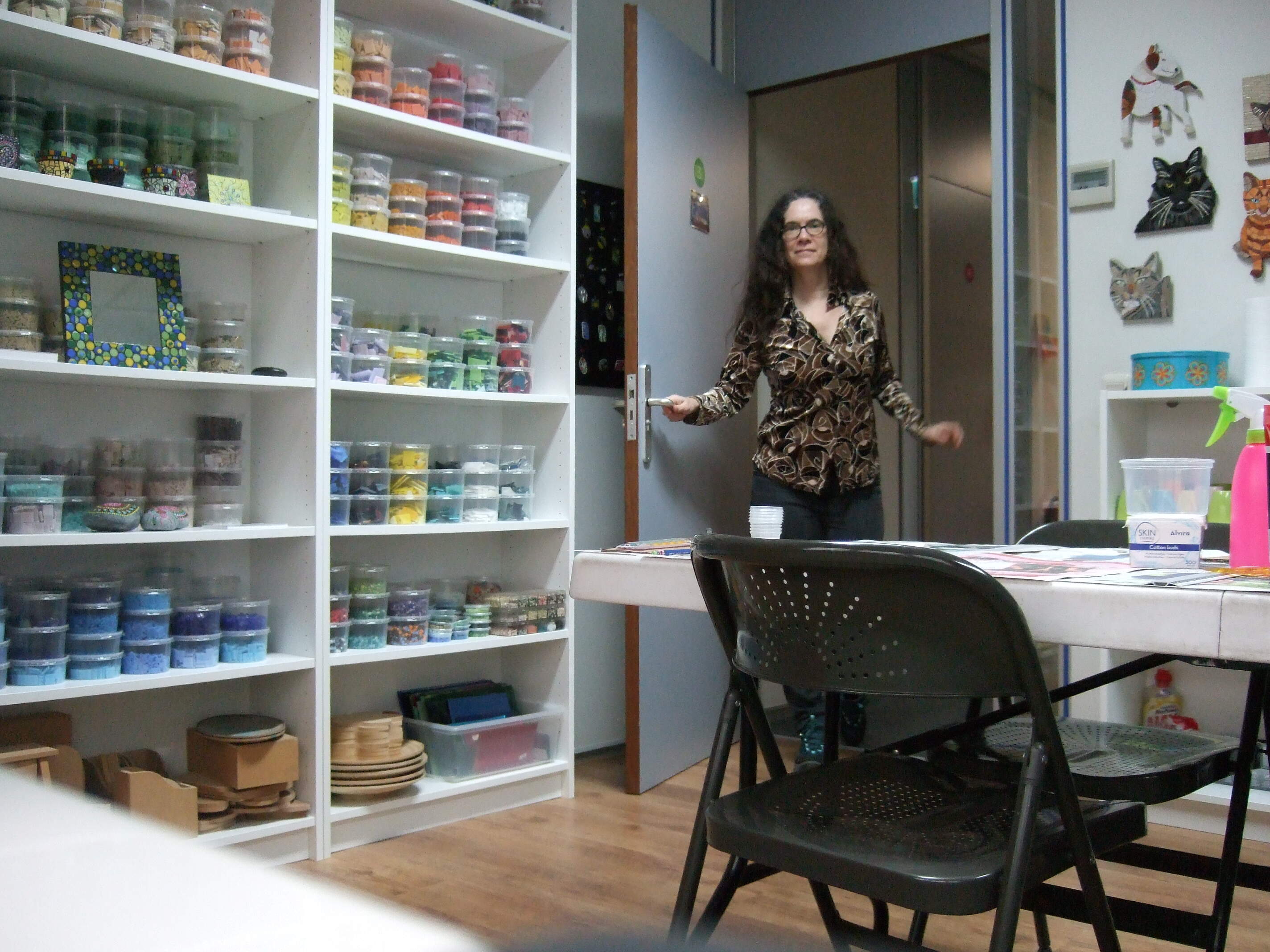Kunstenares Hella Steins komt haar atelier binnen. Het atelier staat vol met mozaiek en dot paintings. De kasten zijn gevuld met kleurrijk mozaiekmateriaal.