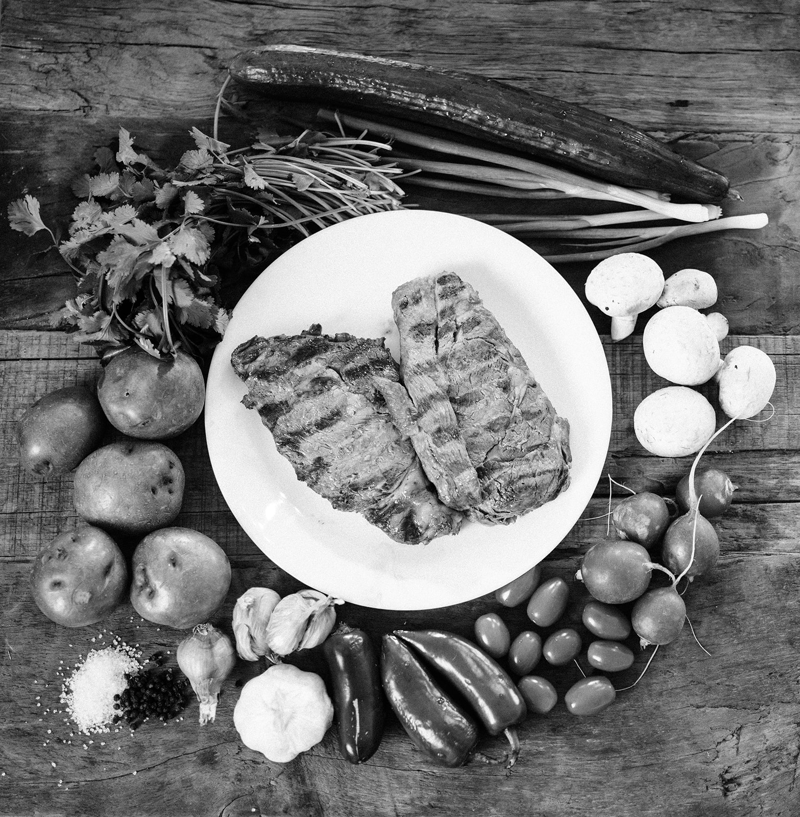A black and white image of a boneless ribeye with grill marks, surrounded by garden vegetables.