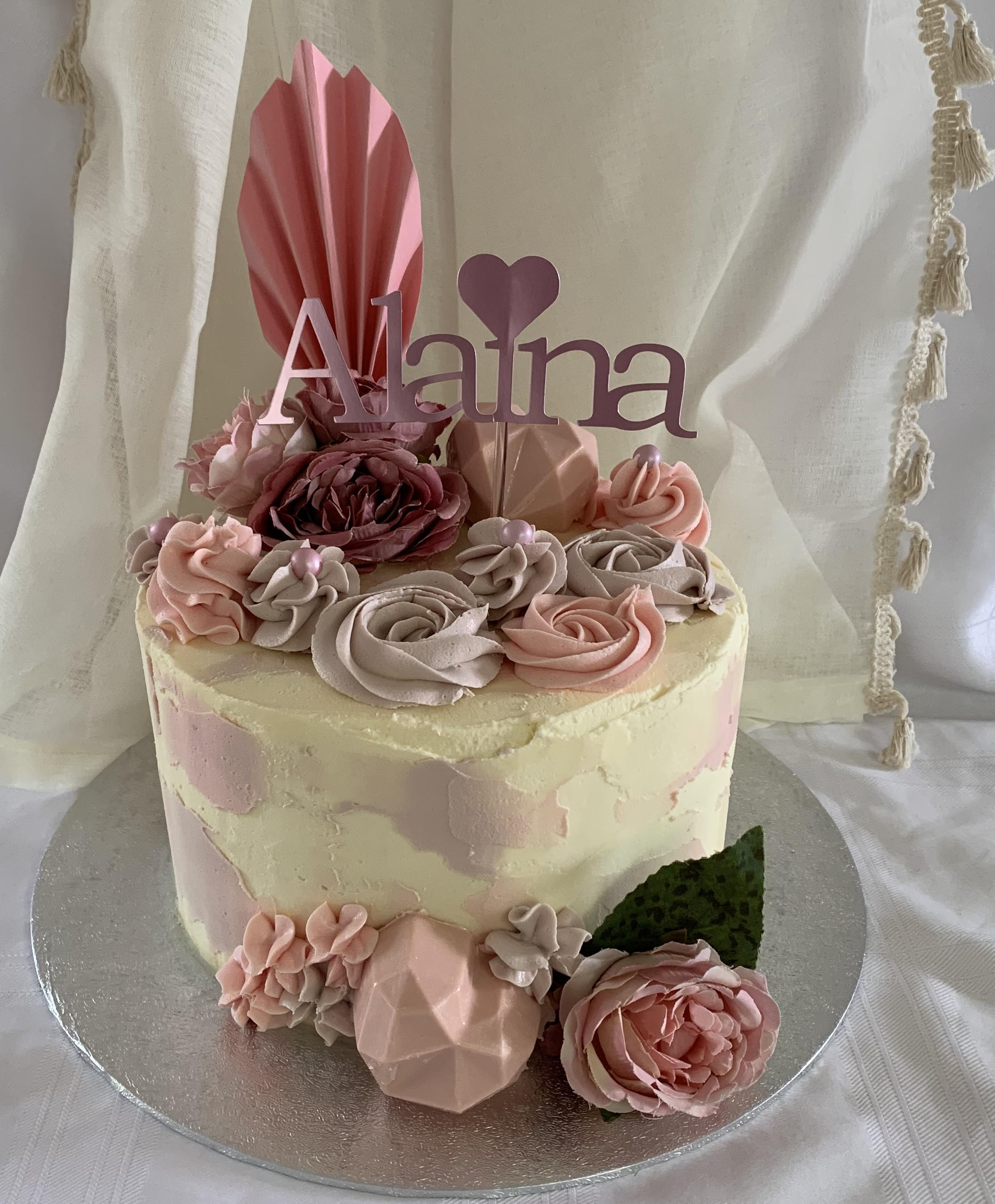 Cake with cream and lilac frosting. There are pink faux flowers, buttercream roses, a chocolate geo heart and a name topper
