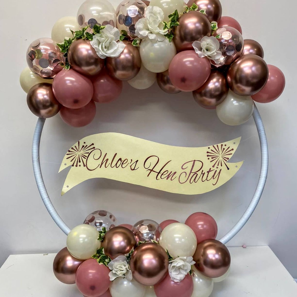 A balloon hoop with personalised banner for Chloes Hen Party in Rose Gold, Cream mini balloons and floral accesories all elegantly styled on a hoop shape