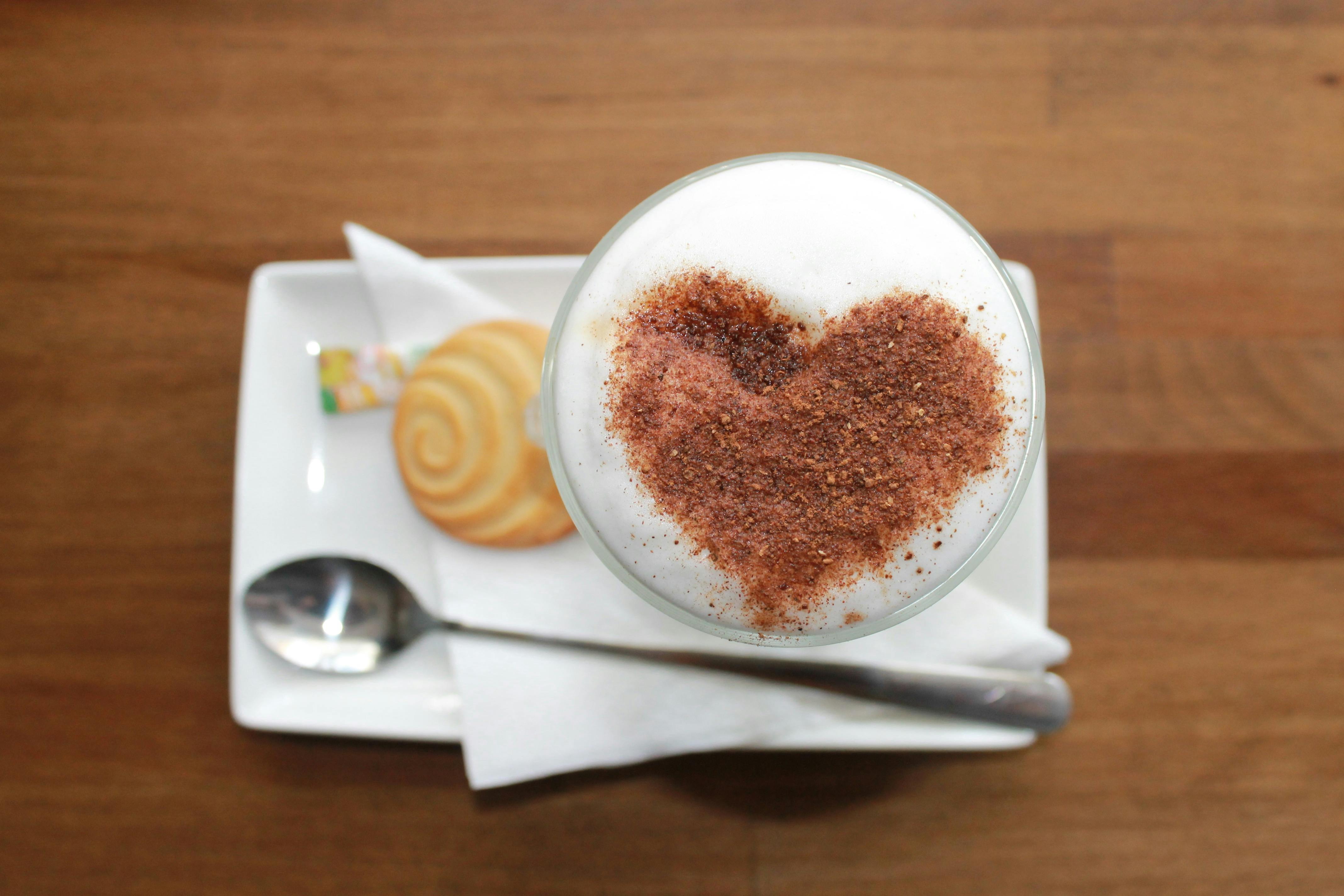 Eine ansprechende Draufsicht auf einen Cappuccino mit herzförmigem Kakaopulvermuster auf der cremigen Schaummütze. Serviert auf einer weißen Untertasse, begleitet von einem spiralförmigen Keks, einem Löffel und einer Serviette, alles auf einem rustikalen Holztisch.